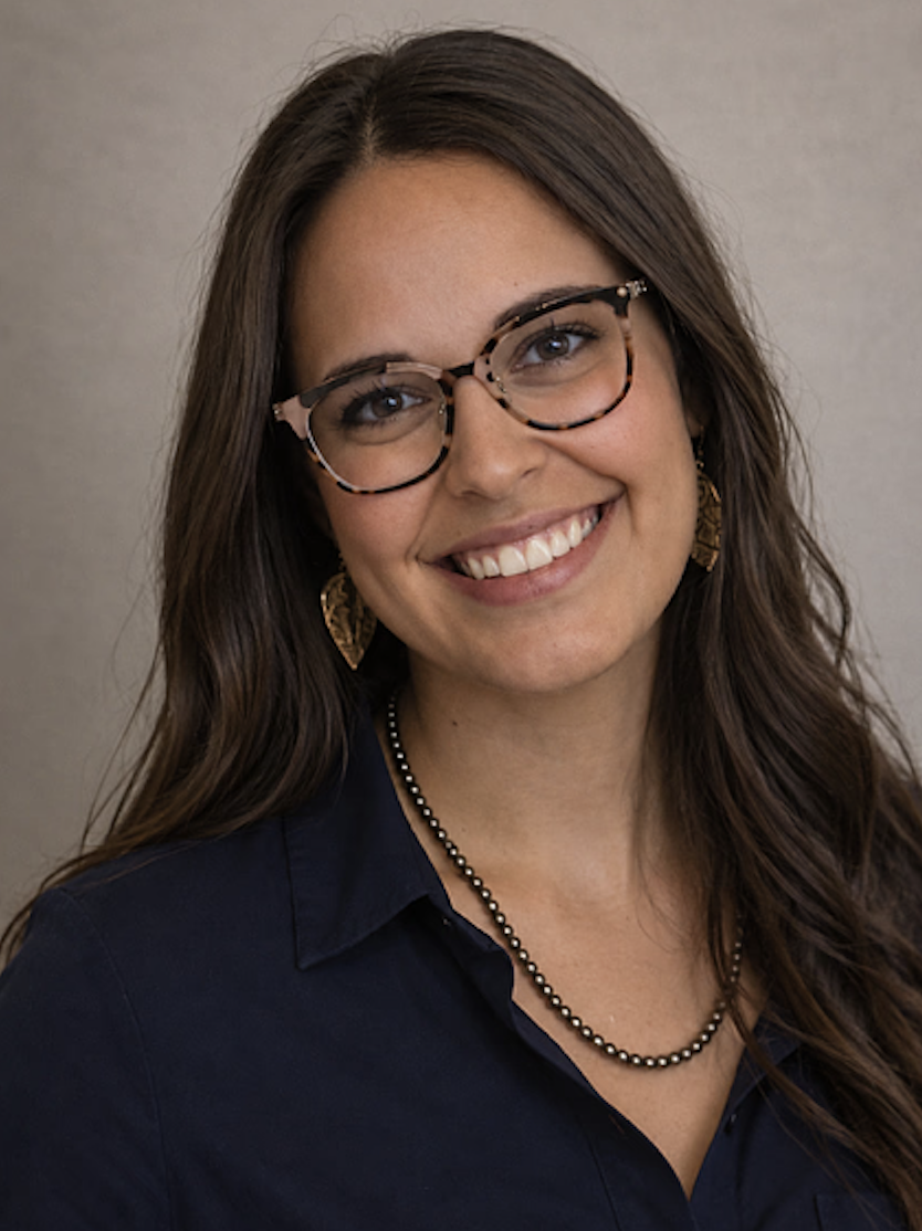 Smiling woman with long dark hair and tortoiseshell glasses, wearing a navy button-down shirt and a beaded necklace.